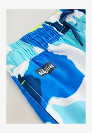 Sinine ja valge mustriga ujumispüksid, millel on elastne vöökoht, string ja must silt, millel on sõna "SUBSCRIBE". Sile kangatekstuur.