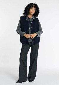 Marineblauer ärmelloser Weste aus strukturiertem Material, großen Knöpfen und oversize Passform, kombiniert mit einem grauen Hemd und dunklen weiten Jeans.