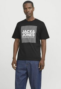 Tricou negru din bumbac, cu un design grafic alb, având modele în zig-zag și textul "JACK & JONES" într-un print pătrat. Guler rotund, mâneci scurte.