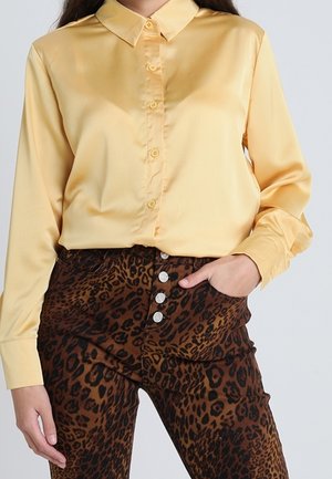 Nainen, jolla on keltainen satiinipaitapusero, joka on laitettu korkeavyötäröisten leopardikuosisten housujen sisään. Housuissa on hopeiset napit.