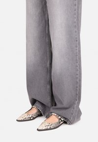 Mocasines puntiagudos con un diseño de estampado de serpiente que presentan una superficie texturizada, complementados por pantalones de mezclilla gris anchos. Suelas negras visibles.