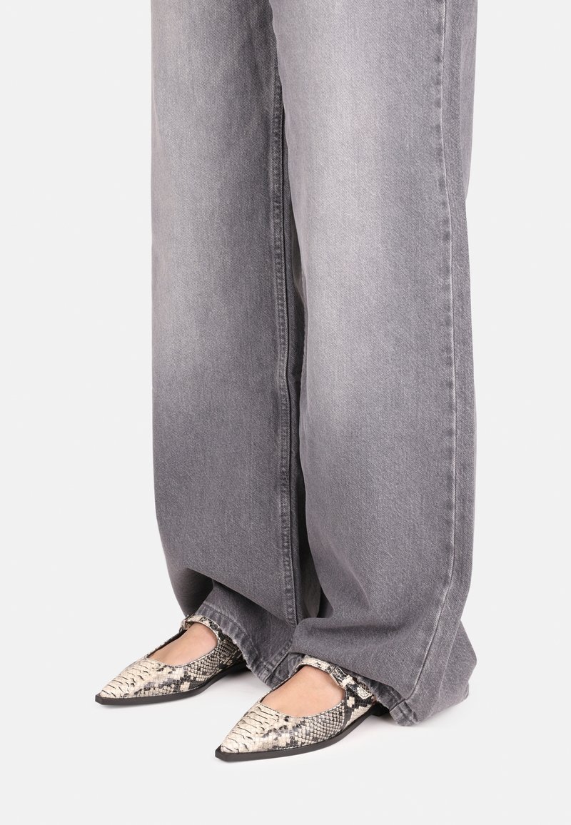 Mocasines puntiagudos con un diseño de estampado de serpiente que presentan una superficie texturizada, complementados por pantalones de mezclilla gris anchos. Suelas negras visibles.