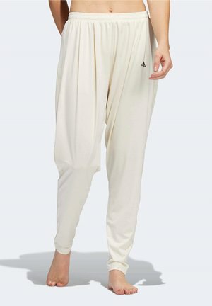 Pantalon de survêtement ample couleur crème Adidas avec taille élastique porté pieds nus sur une personne visible du milieu du torse jusqu’aux pieds.
