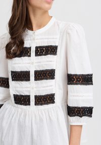 Robe blanche en coton avec des accents en dentelle noire et un devant à boutons. Comprend des manches bouffantes et des panneaux en dentelle horizontaux. Matière en coton douce.