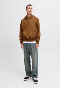 Brązowa bluza z kapturem z przednią kieszenią, zestawiona z jasnoniebieskimi, szerokimi dżinsami z przetarciami oraz czarnymi tenisówkami z białymi dodatkami.