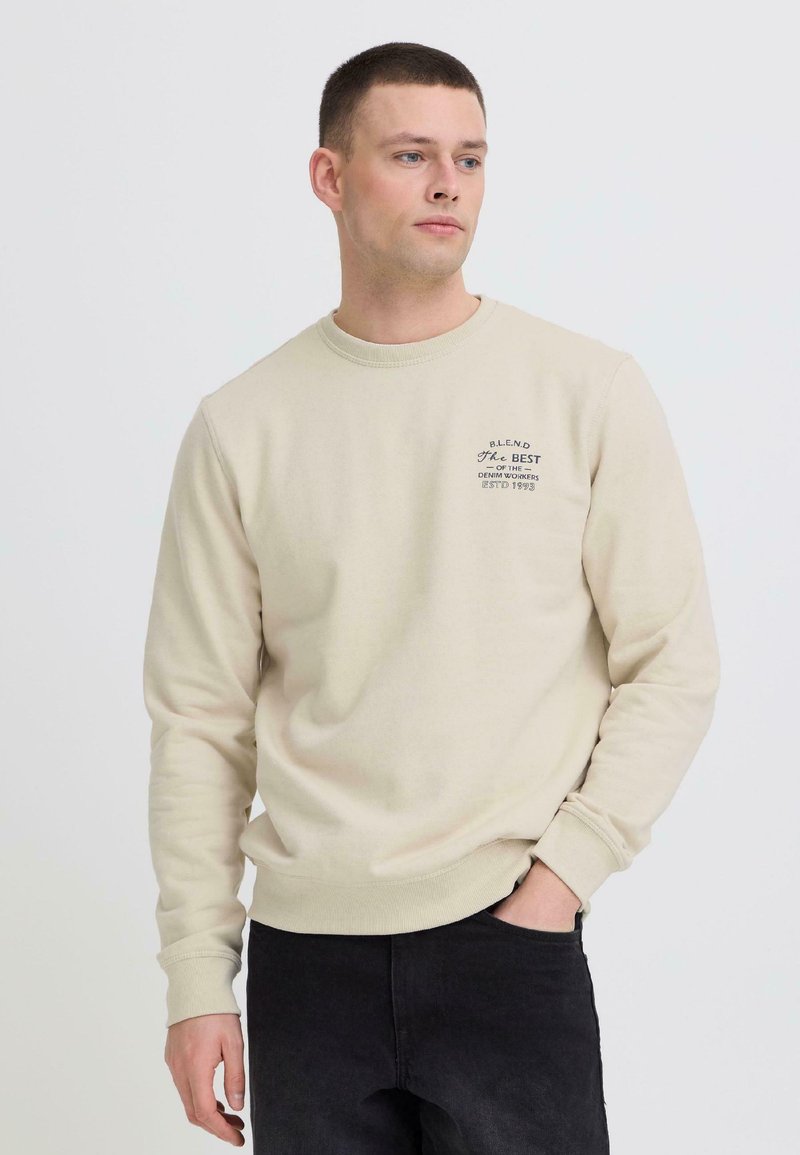 Jeune homme en sweat-shirt beige avec un texte sur la poitrine et un jean noir, debout avec une main dans la poche, regardant légèrement vers la droite.