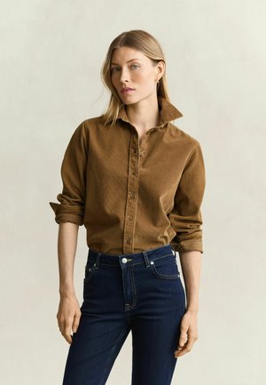 Frau mit blondem Haar, die ein braunes, geknöpftes Hemd mit hochgekrempelten Ärmeln und dunkelblaue, hoch taillierte Jeans vor einem einfarbigen Hintergrund trägt.