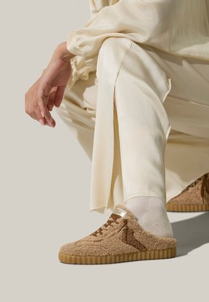 Scarpe marrone chiaro con texture realizzate in materiale morbido, dotate di suola in gomma, con lacci marroni e marchio laterale, posizionate su uno sfondo neutro.