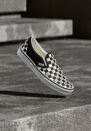 Μαύρο και άσπρο καρό παπούτσι Vans slip-on με μεταλλικά τρουκς που αιωρείται πάνω από μια τσιμεντένια επιφάνεια κοντά σε πέτρινα σκαλοπάτια.