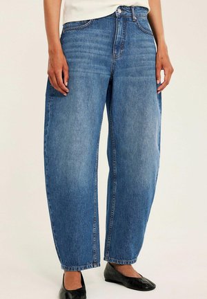 Person trägt locker sitzende blaue Denim-Jeans kombiniert mit schwarzen flachen Schuhen und steht vor einem einfarbigen hellen Hintergrund.