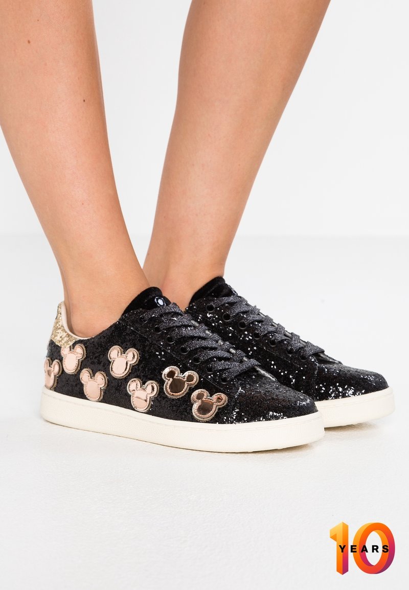 Baskets noires pailletées avec semelles blanches, décorées de multiples formes de têtes de souris dorées et roses sur les côtés, portées pieds nus.