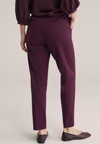 Burgundské kalhoty na míru se slim fit střihem, dvěma zadními kapsami a hladkou strukturou, doplněné o tmavé ploché boty.