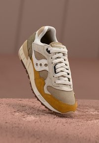 Saucony sneaker with beige mesh, tan suede accents, a white leather logo, and textured cushioning. Features a lace-up design and rubber sole.