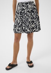 Jupe évasée noire et blanche à motif de feuilles, avec une taille élastique et une longueur au-dessus du genou. Portée avec des tongs noires.