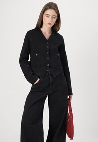 Veste en jean noire avec un col en V, cinq boutons, deux poches poitrine et un design cropped, associée à un pantalon noir à jambes larges.