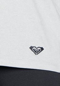 Vaaleanharmaa kangaspaita, jossa on pieni musta kirjailtu sydänlogo helman lähellä, pehmeällä tekstuurilla ja rentolla istuvuudella.