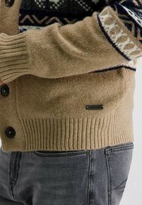 Beige gebreide cardigan met donkere knopen, voorzien van een patronen yoke in marineblauw en wit. Geribde manchetten en zoom, met een discreet logo-label.
