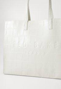 White crocodile-embossed tote bag with smooth leather, featuring two handles and embossed logo "TED BAKER LONDON." Structured rectangular shape.