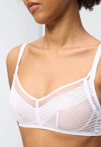 Γυναίκα που φοράει λευκό δίχτυ με γεωμετρικά σχέδια, εμφανίζοντας τον λαιμό και τους ώμους της σε απλό φωτεινό φόντο.