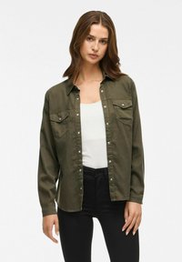 Camicia verde oliva con bottoni in cotone, dotata di due tasche sul petto e bottoni a pressione bianchi, abbinata a una maglietta bianca e jeans neri.