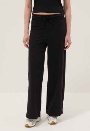Mujer con pantalones negros de pierna ancha con cintura elástica con cordón y zapatillas blancas, de pie sobre fondo liso.