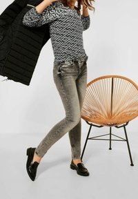 Vadderad svart jacka, geometriskt mönstrad grå topp, grå tajta jeans och blanka svarta loafers; en rottingsstol i bakgrunden.