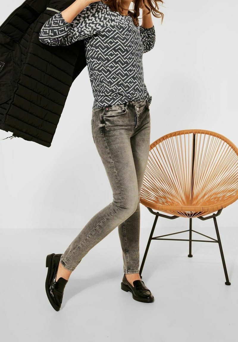 Vadderad svart jacka, geometriskt mönstrad grå topp, grå tajta jeans och blanka svarta loafers; en rottingsstol i bakgrunden.