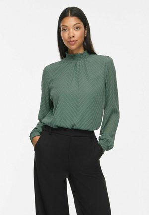 Femme portant un chemisier vert texturé à manches longues avec un col volanté et un pantalon noir taille haute, debout les mains dans les poches.