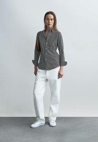 Πουκάμισο με κουμπιά με κάθετες ασπρόμαυρες ρίγες, σε συνδυασμό με άνετο λευκό παντελόνι και λευκά αθλητικά παπούτσια· κρατά μια καφέ τσάντα.