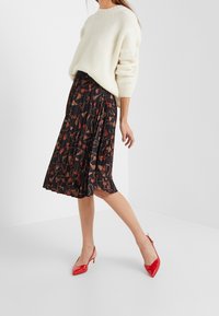 Svart plisserad kjol med ett blommönster i röd och beige, kombinerad med en krämfärgad stickad tröja och röda spetsiga klackskor.