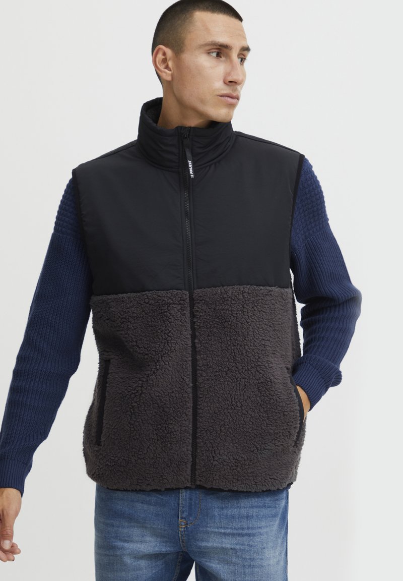 Fleece mellény fekete felső résszel és sötétszürke bolyhos alsó résszel, álló nyakkal és texturált kék ujjakkal, cipzáras elülső résszel és zsebekkel.
