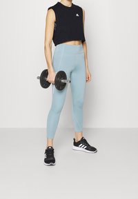 Černý bezrukávový crop top, světle modré legíny s vysokým pasem, černé sportovní boty a činka v ruce, stojící na jednobarevném pozadí.