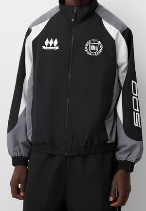 Zwart, grijs en wit sportjack met rits, met een "MRA" wapen, "Original Alumni" logo en de tekst "500" op de mouw, gedragen door een staande persoon.