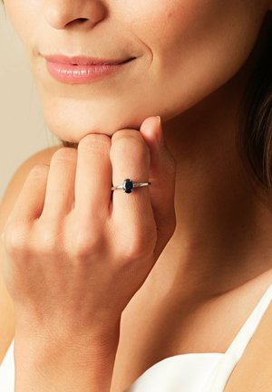 Frau, die einen Silberring mit einem dunkelblauen ovalen Stein an ihrem Finger trägt, die Hand sanft an ihrem Kinn berührend, mit einem sanften Lächeln im Gesicht.