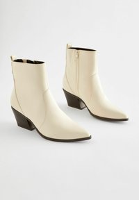 Bottines en cuir crème avec un bout pointu et un talon empilé. Présentent une fermeture éclair sur le côté et une couture simple pour un détail supplémentaire.