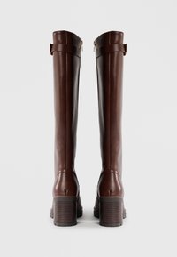 Bottes hautes en cuir marron avec un design structuré, un talon épais en bloc et des accents de sangle en haut. Texture lisse, détails minimaux.