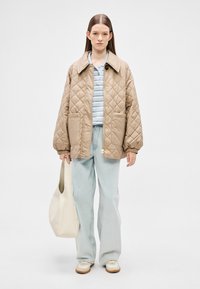 Μπεζ μπουφάν με καπιτονέ και μεγάλο γιακά, σε συνδυασμό με γαλάζιο πουκάμισο με ρίγες και ανοιχτό μπλε παντελόνι καμπάνα. Κρατάει λευκή τσάντα tote.