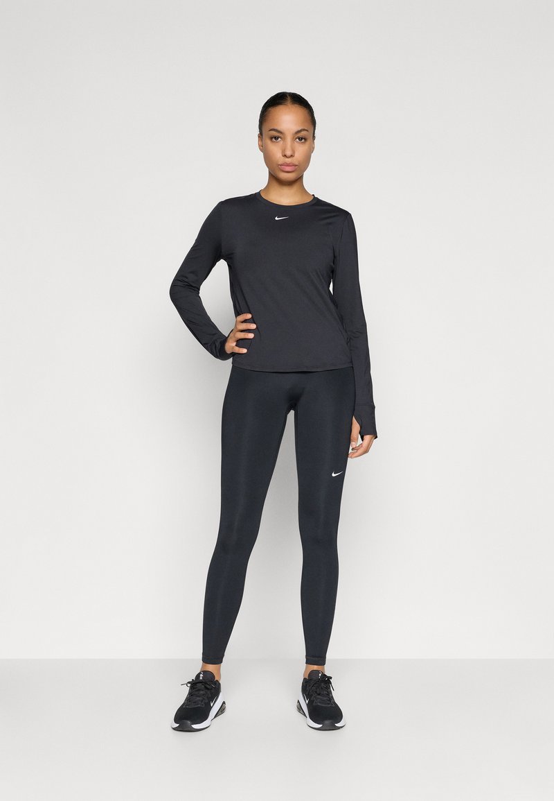 Zwarte sportieve top met lange mouwen en zwarte leggings, beide met een klein wit Nike-logo, gecombineerd met zwarte sneakers. Soepele, aansluitende stof.