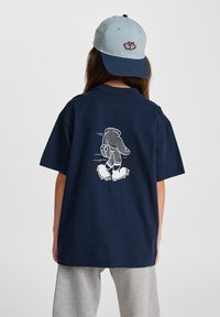 Marinblå bomullst-shirt med en grafik av en karaktär på rullskridskor i vitt på baksidan, tillsammans med grå mjukisbyxor och en ljusblå keps.