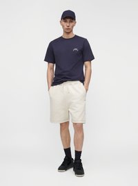 Jeune homme debout, les mains dans les poches, portant une casquette et un t-shirt bleu marine, un short beige, des chaussettes noires et des baskets noires sur fond blanc uni.