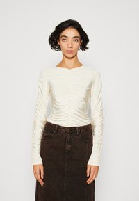Ruched langærmet top i cremet satin stof med rund halsudskæring og tætsiddende design, parret med en højtsiddende brun denimnederdel.