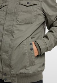 Chaqueta verde oliva de un tejido resistente, con bolsillos en el pecho con cierre de botón, puños acanalados y cierre de cremallera. Se visible un bolsillo lateral.