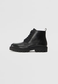 Botas negras de cuero hasta el tobillo con punta redonda, cordones en la parte delantera y suela de goma texturizada. Incluye una pestaña en el talón para ponerlas y quitarlas fácilmente.