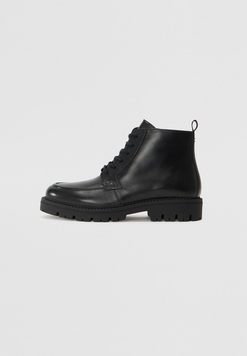 Botas negras de cuero hasta el tobillo con punta redonda, cordones en la parte delantera y suela de goma texturizada. Incluye una pestaña en el talón para ponerlas y quitarlas fácilmente.