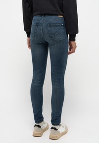 Jeans ajustados de mezclilla azul oscuro, que presentan dos bolsillos traseros con costura curvada y una etiqueta color beige en la cinturilla.