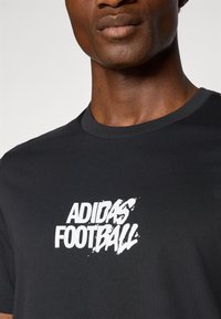 Crna pamučna majica s bijelim grafičkim logom "ADIDAS FOOTBALL". Okrugli izrez s običnom, glatkom teksturom. Minimalistički dizajn.