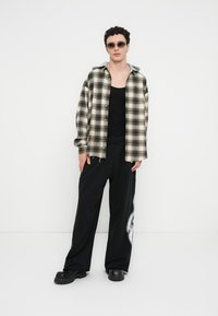 Chemise à carreaux noire et beige avec fermeture à boutons, associée à un pantalon noir ample orné d'un imprimé graphique. Le mannequin porte des lunettes de soleil et un débardeur noir.