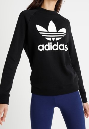 Femme portant un sweat-shirt Adidas noir avec logo blanc et des leggings de sport bleus avec des bandes blanches sur les côtés.