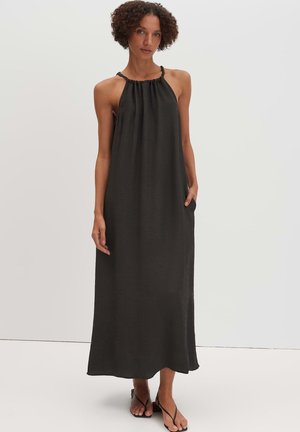 Frau, die ein ärmelloses, schwarzes Midi-Kleid mit Neckholder und Taschen sowie schwarze Riemchensandalen trägt, steht vor einem schlichten Hintergrund.