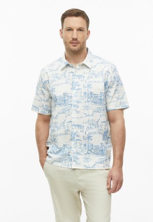 Homme aux cheveux châtain clair portant une chemise blanche à manches courtes avec des imprimés bleus représentant des paysages et un pantalon beige clair, debout devant un fond blanc.